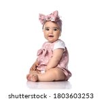 Small photo of Excited amazed barefooted Infant baby toddler in polka-dot dress and headband with bow sits on the floor looking at camera with her mouth open. Happy infancy and babyhood concept