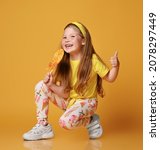Small photo of Frolic laughing red-haired seven years old kid girl in yellow t-shirt and leggings with lollipop ice-cream pattern, print sits squatting, holding big lollipop and showing thumbs up sign, gesture