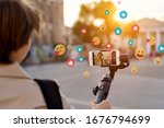 Small photo of Teen girl blogger vlogger record vlog streaming video hold phone on selfie stick in urban city. Young female vlogger shoot social media blog on smartphone get likes emoji, over shoulder closeup view.