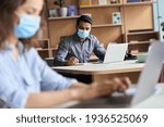 Small photo of Indian professional business man or student wearing medical face mask working, studying on laptop among diverse people sitting at table in modern office coworking space keeping safe social distance.