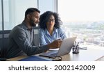 Small photo of Indian male ceo executive manager mentor giving consultation on financial operations to female African American colleague intern using laptop sitting in modern office near panoramic window.