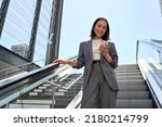Smiling young Asian business woman wearing suit standing on urban escalator using applications on cell phone, reading news on smartphone, fast connection, checking mobile apps outdoors.