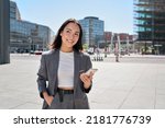 Young smiling elegant Asian busy business woman leader wearing suit standing in big city using cell phone platform applications. Smiling woman holding smartphone walking on street outdoors.