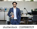 Small photo of Smiling elegant young indian business man professional manager, arab company employee, eastern businessman executive financial banker wearing suit standing in modern office holding laptop, portrait.
