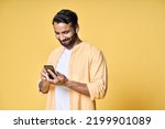 Happy smiling indian ethnic man wearing earbuds using smartphone looking at cell phone watching videos, having virtual mobile chat video call, playing game isolated yellow background.