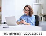 Young happy business woman, smiling pretty professional businesswoman worker looking at smartphone using cellphone mobile technology working at home or in office checking cell phone sitting at desk.