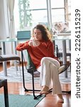 Small photo of Young adult smiling pretty woman sitting on chair holding cup drinking tea or coffee relaxing at home. Happy calm lady enjoying warm hot drink with mug in hands daydreaming in cozy morning. Vertical