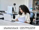 Small photo of Young latin business woman manager accounting analyst checking bills, analyzing sales statistics management, taxes financial data documents or marketing report papers working in office using laptop.