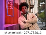 Small photo of Confident cool African American hipster guy standing at city street near neon light sign. Rebel gen z male model looking at camera outside advertising young generation culture life entertainment.