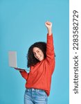 Small photo of Young happy latin woman winner holding laptop isolated on blue background. Excited lucky female student using computer winning online celebrating success admission with yes gesture concept.