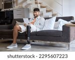 Small photo of Happy young man with disability leg prosthesis sitting on sofa at home using digital tablet working or elearning, browsing web, searching online. People with amputation disabilities everyday life.