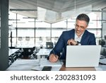 Small photo of Middle aged smiling professional business man global company executive ceo manager or lawyer wearing suit sitting at desk in modern office working on laptop computer and writing notes, copy space.