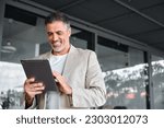 Smiling elegant mid aged business man wearing suit standing outside office holding digital tablet. Mature businessman professional using fintech device working on modern technology gadget.