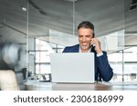 Happy middle aged business man talking on mobile phone working on laptop in office. Professional businessman executive wearing suit making call on cell using computer communicating with client.