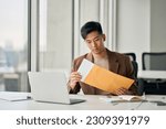 Small photo of Young busy Asian professional business man manager opening envelope delivery receiving business mail letter holding paper documents bank statements, invoice file or bill sitting at desk in office.