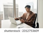 Young busy serious Asian Japanese business man worker executive holding cellphone using mobile phone looking at laptop checking financial market online in app working in corporate office at desk.