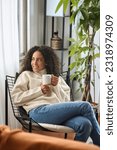 Small photo of Young adult smiling pretty latin woman sitting on chair holding cup drinking tea or coffee relaxing at home. Happy calm lady enjoying warm hot drink with mug in hands daydreaming. Vertical