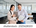 Two happy professional business people team Asian woman and Latin man workers working using digital tablet tech discussing financial market data standing at corporate office meeting.
