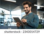 Happy young Latin business man executive holding pad computer at work. Male professional employee using digital tablet fintech device standing in office checking financial online market data.