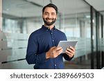 Small photo of Smiling professional Indian businessman entrepreneur using tab computer working standing in office. Confident business man holding digital tablet tech device working, looking at camera. Portrait.