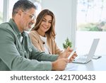 Small photo of Happy middle aged mature couple using laptop counting taxes refund receipts to save money at home. Old man and woman paying bills online planning financial budget savings calculating payment at table.