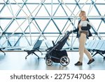 Small photo of Mother carying his infant baby boy child, pushing stroller at airport departure terminal moving to boarding gates to board an airplane. Family travel with baby concept