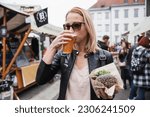 Beautiful young woman holding delicious organic salmon vegetarian burger and drinking homebrewed IPA beer on open air beer an burger urban street food festival in Ljubljana, Slovenia