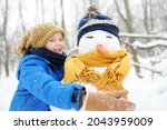 Little boy building snowman in snowy park. Child embracing snowman wearing hat and scarf. Active outdoors leisure with family with children in winter. Kid during stroll in a snowy winter park
