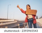 Woman is hitchhiking on roadside trying to stop car. She is holding cardboard with inscription.