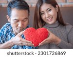Small photo of Lover couple happy in love relaxing on sofa looking in eye smiling teasing with big red heart shape pillow in valentine day honeymoon. teasing teasing with red heart pillow.