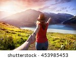 Woman in orange t-shirt and hat holding man by hand and going to the lake in the mountains at glowing sunrise 