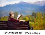 Woman in red checked shirt and hat sitting on the wooden bench and reading book at autumn forest and mountains background