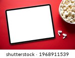 Tablet with empty white screen with wireless earphones and bowl of popcorn next to it. Mockup for video or movie screenshot.
