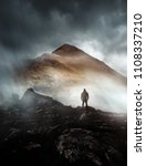 Small photo of A person hiking looks onwards at a mountain shrouded in mist and clouds with the peak visible. Scenic landscape photo composite.