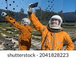 Happy astronaut wearing an orange spacesuit and space helmet holding mobile phone and taking selfie outdoors. Cheerful cosmonaut making a photo of himself standing against futuristic mirror wall