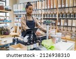 Small photo of African american saleswoman or small business owner weighs some kraft paper package on scales at sustainable small local business. Young afro woman at cashdesk with scales in local zero-waste grocery