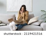 Small photo of Happy young woman sit on couch in living room unpack cardboard box buying goods on Internet, smiling excited millennial girl open carton parcel order, shopping online, good delivery concept