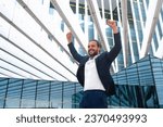 Excited businessman in suit celebrating victory arms raised up. Hispanic male business person winner pose. Best deal ever. Business man expressing positivity standing outdoors office building