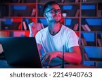 Positive emotions. Man in glasses and white shirt is sitting by the laptop in dark room with neon lighting.