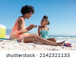 Small photo of Happy african american woman applying suntan lotion on daughter while sitting at beach on sunny day. unaltered, family, lifestyle, togetherness, enjoyment and holiday concept.