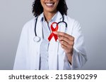 Midsection of smiling african american mid adult female doctor holding red aids awareness ribbon. white background, hiv, aids, doctor, medical, disease, awareness, support and healthcare concept.