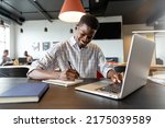 Smiling young african american businessman writing in diary and using laptop in creative office. Unaltered, creative business, workplace, occupation, wireless technology.
