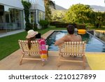 Small photo of Rear view of a Caucasian couple wearing beachwear sitting on chairs sunbathing in a garden beside a swimming pool, the woman wearing a sun hat and using a laptop
