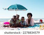 Small photo of Smiling african american parents and children lying on towels at sandy beach under clear sky. Copy space, unaltered, family, together, parasol, picnic, nature, vacation, enjoyment, relaxing, summer.