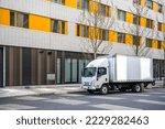 Small photo of Small rig over cab white semi truck with long box trailer making local commercial delivery at urban city with multilevel residential apartments buildings standing on the street for upload cargo
