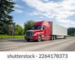 Small photo of Red big rig popular professional reliable bonnet long haul semi truck transporting commercial cargo in dry van semi trailer moving on the straight wide highway with green trees on the background