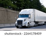 Elegant powerful white modern bonnet big rig long haul semi truck transporting commercial cargo in dry van semi trailer on the road with concrete side wall and trees on the hill