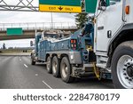 Small photo of Powerful blue big rig heavy duty towing semi truck tractor with lifting boom and winch tow broken semi truck driving on the wide highway road under the bridge