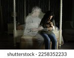 A sad woman clutching a photo of herself and her dead husband is being consoled by his ghost as she sits by her bed crying in her dimly lit bedroom. Life after death, heaven and the afterlife.