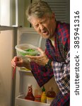 Small photo of A man hunting for a midnight snack in the refrigerator and has found a container with stinking spoiled rotten food which has made his face twisted with his wretched disgust.